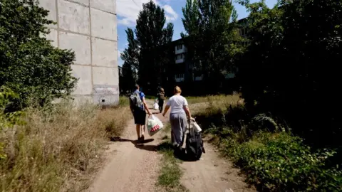 Three people walk down a dirt path past a building and piles of weeds, carrying large bags