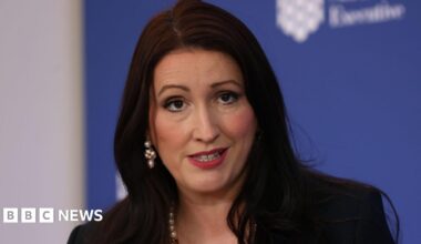 Northern Ireland Deputy First Minister Emma-Little Pengelly is speaking during a press conference. We can see the tops of her shoulders and head. She has shoulder-length dark hair and is wearing a pearl necklace and pearl earrings. She is wearing a dark jacket. In the background is a blue board with the logo of the Northern Ireland Executive