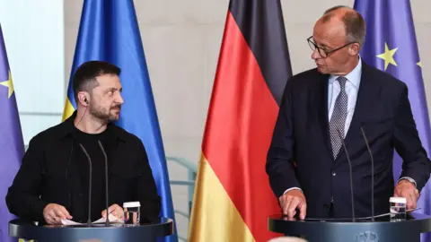 EPA Volodymyr Zelensky and the German chancellor stand in front of two lecterns looking at each other. The flags of Ukraine and Germany are hanging behind them.