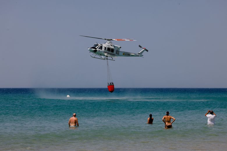 People watch as a helicopter draws water at Atlanterra Beach on August 12, 2025, a day after many people, including locals and tourists, were evacuated from Tarifa due to wildfires.