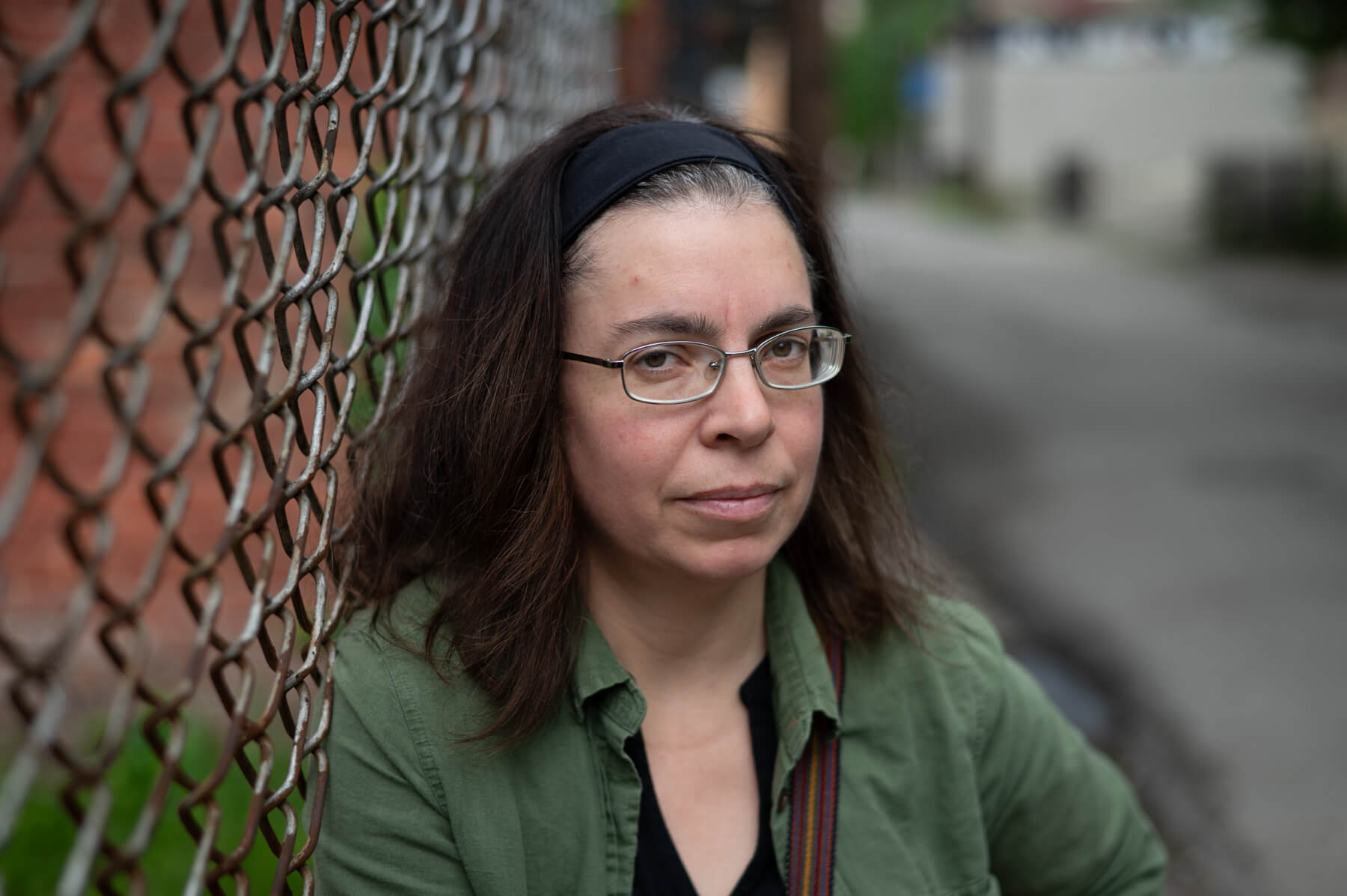 A woman wearing glasses and a green shirt leans against a chain-link fence on an urban street.