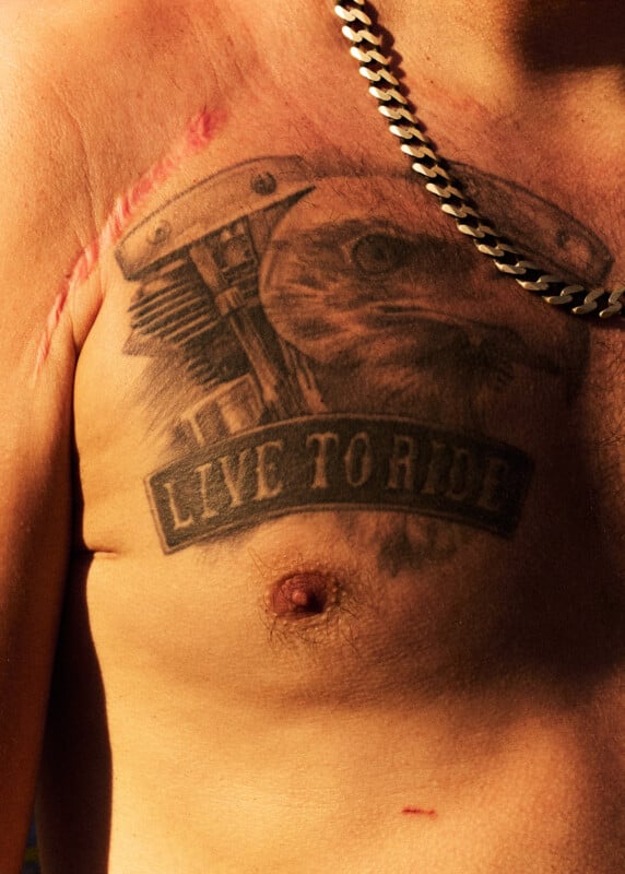 A close-up of a man's chest showing a tattoo of an eagle and engine parts with the words "LIVE TO RIDE," a noticeable scar above it, and a metal chain necklace around his neck.