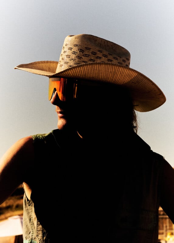 A person wearing a wide-brimmed straw hat and reflective sunglasses stands outdoors in strong sunlight, casting deep shadows across their face and upper body.