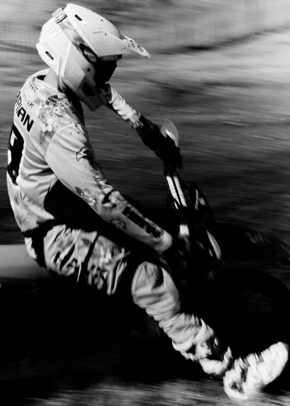 A motocross rider in full gear and helmet is captured mid-action riding a dirt bike, blurred in black and white, emphasizing motion and speed.