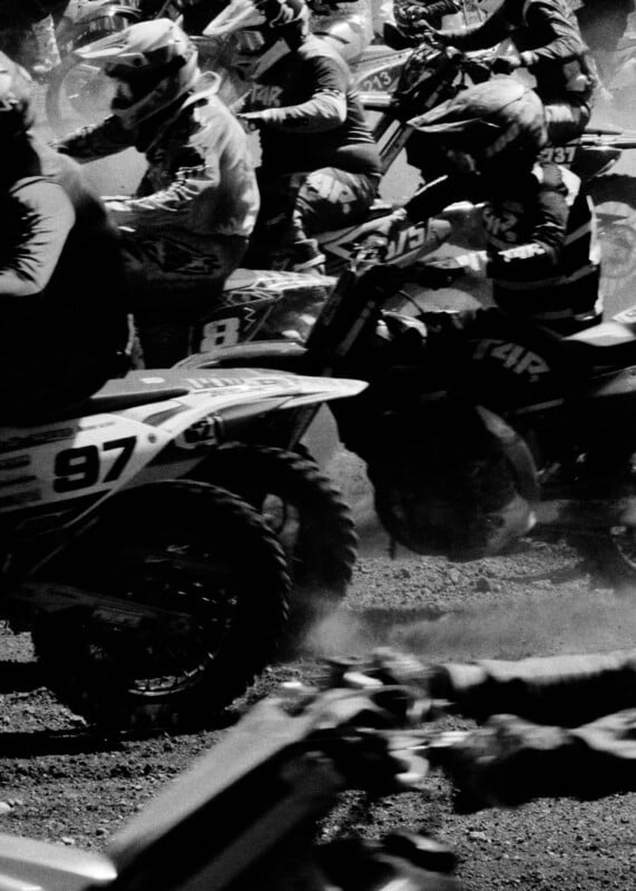 A group of motocross riders wearing helmets race closely together on dirt bikes, kicking up dust as they speed through a turn. The image is in black and white, capturing the intensity of the moment.