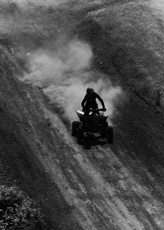 A person rides an ATV up a dirt hill, kicking up a cloud of dust behind them. The scene is in black and white, emphasizing the texture of the dirt and the dramatic motion.