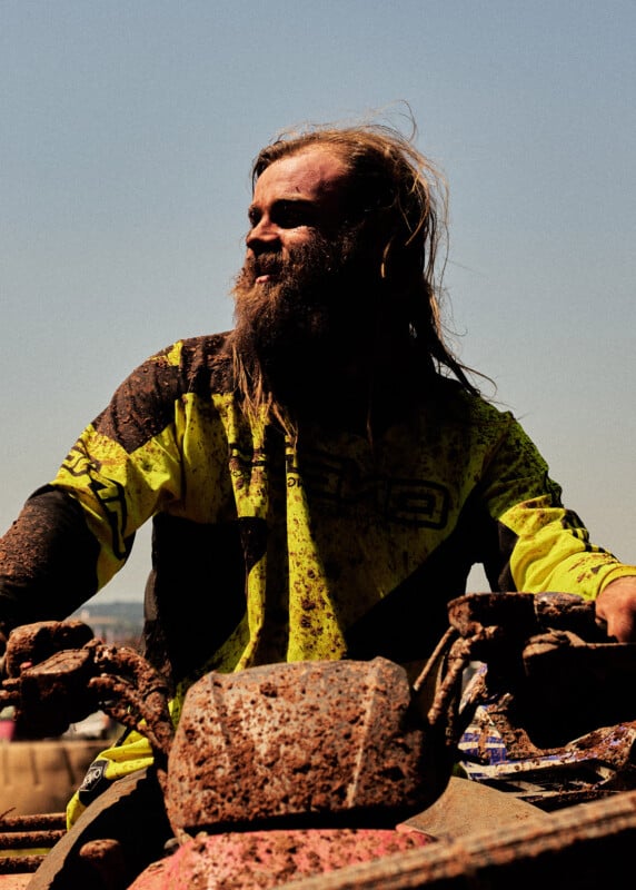 A bearded man wearing a yellow and black jersey, covered in mud, rides an ATV under a clear blue sky. His hair is messy, and he appears focused, gripping the muddy handlebars.