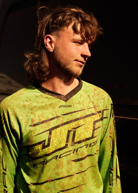 A young man with a mullet hairstyle wears a bright green and black racing jersey. He stands in sunlight, looking to the side, with dirt stains visible on his shirt. The background is dark and shadowed.