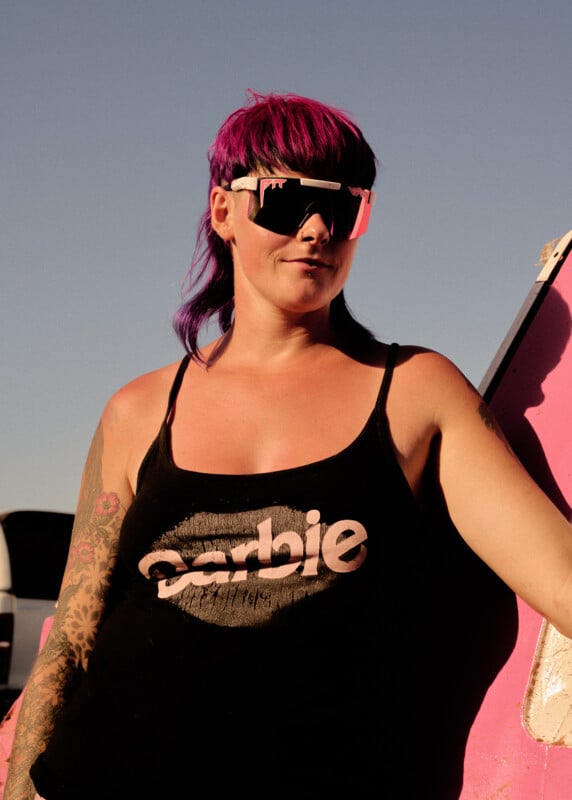 A woman with bright purple hair and large reflective sunglasses wears a black tank top with the word "Barbie" on it, standing outdoors in sunlight next to a pink vehicle.