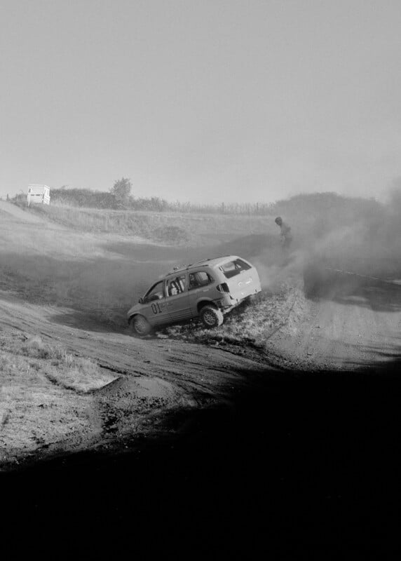 A small car with the number 01 skids off a dirt road, kicking up dust, while a person stands nearby. The scene is outdoors with grassy terrain and a distant vehicle visible. The image is in black and white.
