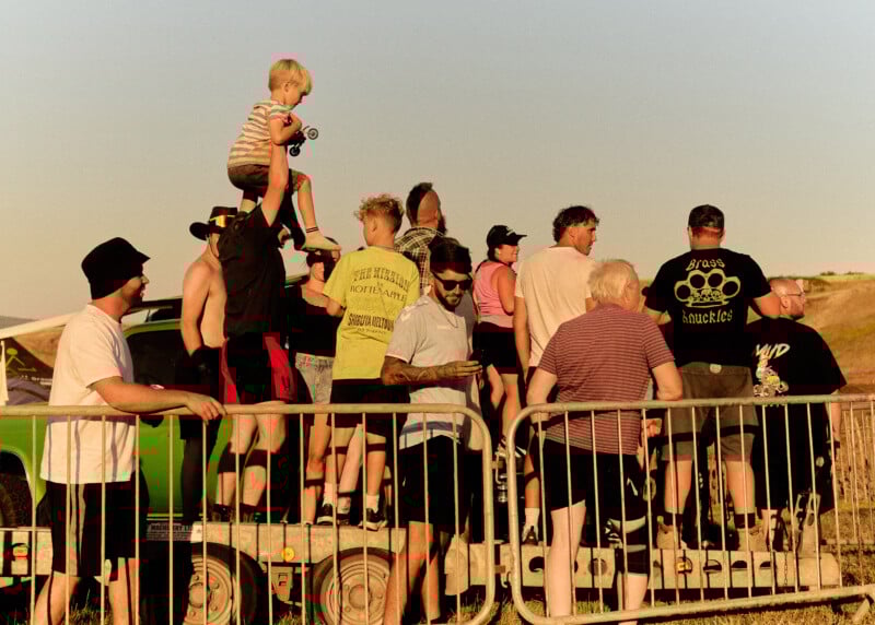 A group of people, including a child on someone’s shoulders, stand on a trailer behind a metal barrier, watching an outdoor event in warm, golden sunlight. The scene suggests a casual, lively gathering.