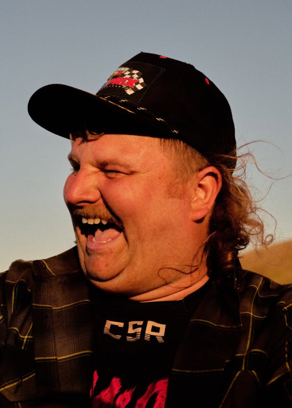 A man with a mustache wearing a black cap and plaid shirt laughs outdoors under a clear sky, with sunlight highlighting his face and hair.