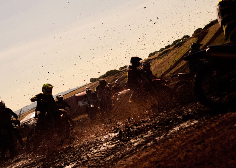 Motorcyclists race through muddy terrain, kicking up dirt and debris. The sun casts a warm light, creating dramatic silhouettes of the riders against the sky and hilly landscape in the background.