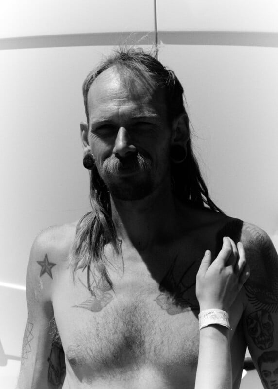 A shirtless man with tattoos, piercings, and long hair stands in sunlight. A person's arm with a hospital wristband rests on his shoulder. The background is plain and bright.