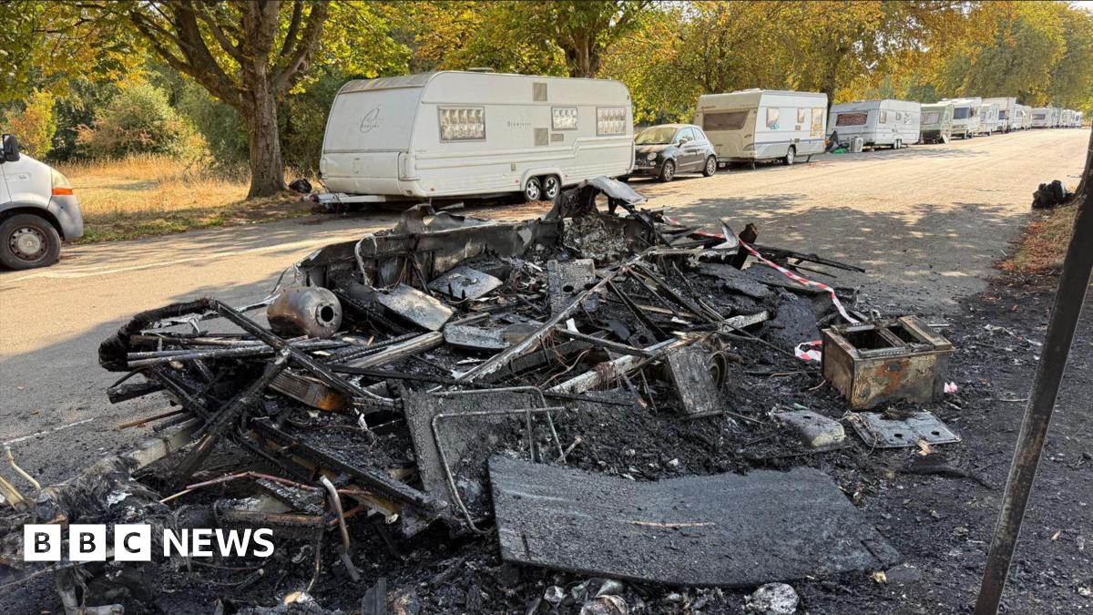 The burnt and charred remains of a caravan on a street running through the Downs. The vehicle has almost been burnt to the ground and there are some personal belongings laying scattered on the burnt grass beside it. There is red and white cordon tape flapping in the breeze. In the background, there are white caravans parked on the other side of the road for as far as the eye can see.