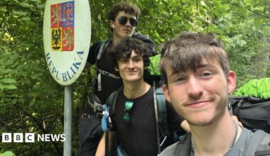 Kyle Spring (L), James McCarthy-Hill, and Adam Mitchell (R) wearing large back packs and posing for a selfie beside the Czech Republic border sign in front of lots of trees.
