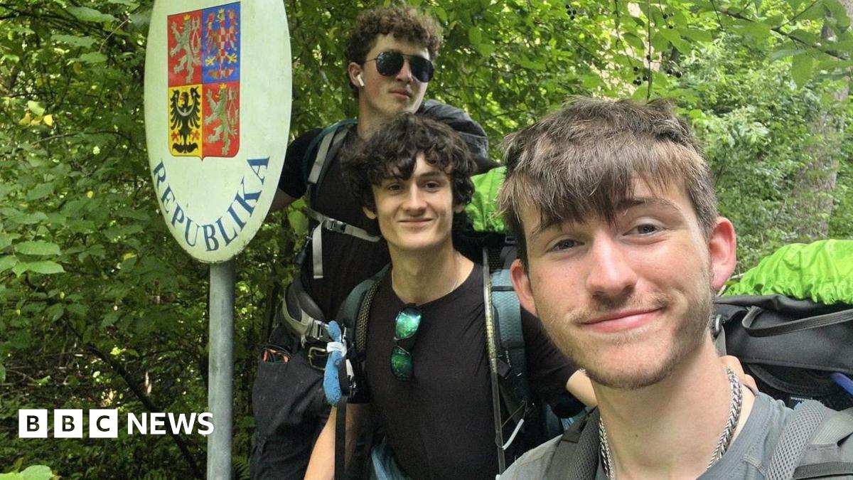 Kyle Spring (L), James McCarthy-Hill, and Adam Mitchell (R) wearing large back packs and posing for a selfie beside the Czech Republic border sign in front of lots of trees.