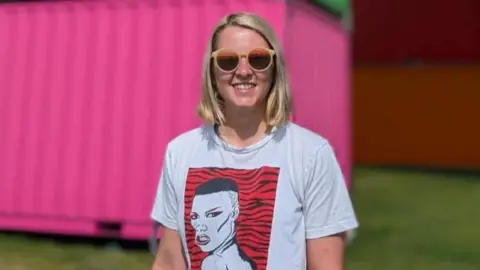 Cissy Badley Cissy Badley. She has short blonde hair and is wearing brown sunglasses. She is smiling at the camera. A bright pink wall can be seen behind her.