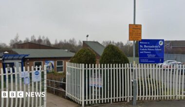 A view of a school with white gates outside it. A blue sign saying 'Welcome to St Bernadette's Catholic Primary School and Nursery' is visible, as is a yellow 'No stopping' sign which has times on it.