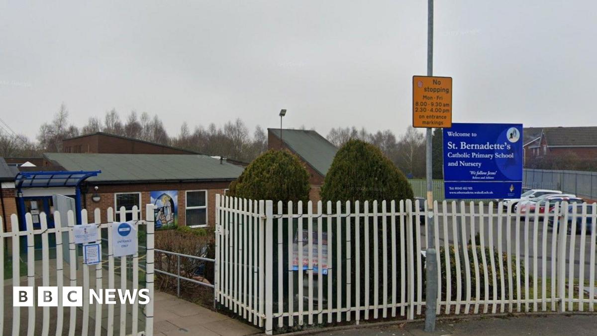 A view of a school with white gates outside it. A blue sign saying 'Welcome to St Bernadette's Catholic Primary School and Nursery' is visible, as is a yellow 'No stopping' sign which has times on it.