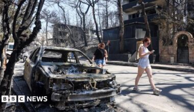 Women in shorts walk past a fully charred and burnt car ion a street surrounded by charred trees