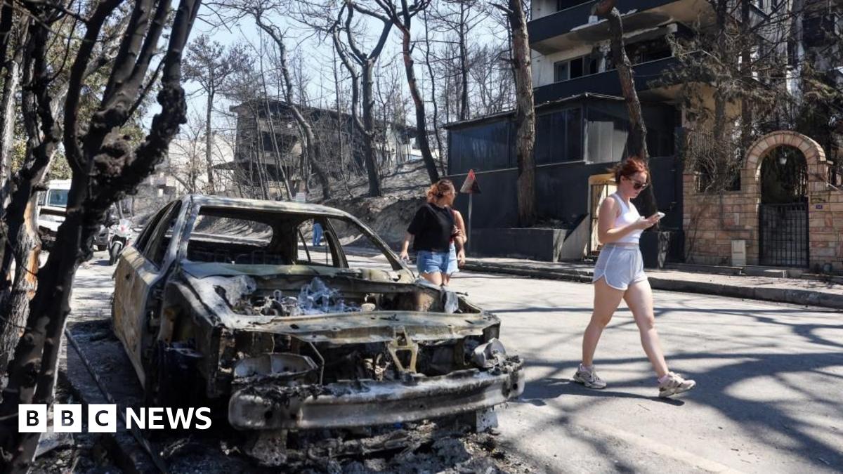 Women in shorts walk past a fully charred and burnt car ion a street surrounded by charred trees