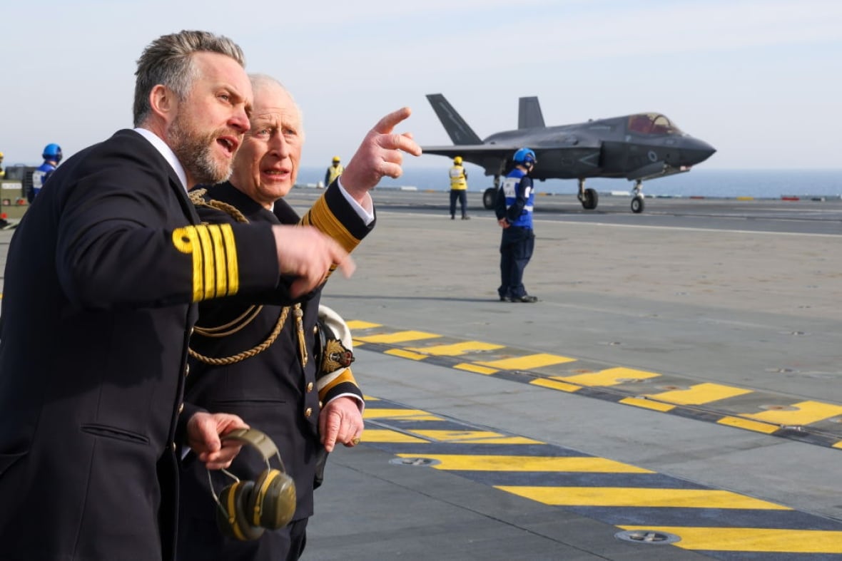 Two people standing close together on the deck of an aircraft carrier point into the distance. An aircraft sits on the deck behind them.