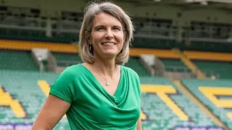 Northampton Saints RUFC Julia Chapman in a green dress. She has light coloured hair cut just above her shoulders and is standing against the backdrop of Northampton Saints stadium.