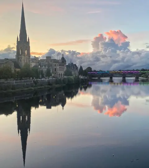 Mark Macleod A view of Perth from across the Tay in the evening - the sky is a light blue with clouds tinged in a pink light. The bridge in the distance is lit in pastel colours adding to the magical feel