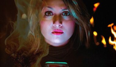 Woman looks directly at the screen. Darkness is behind with some fire. Fierce look.