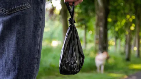 Getty Images A person in blue jeans holding a full black poo bag. A larger cream-coloured dog is blurred in the distance. The background is a green wooded area and is blurred.