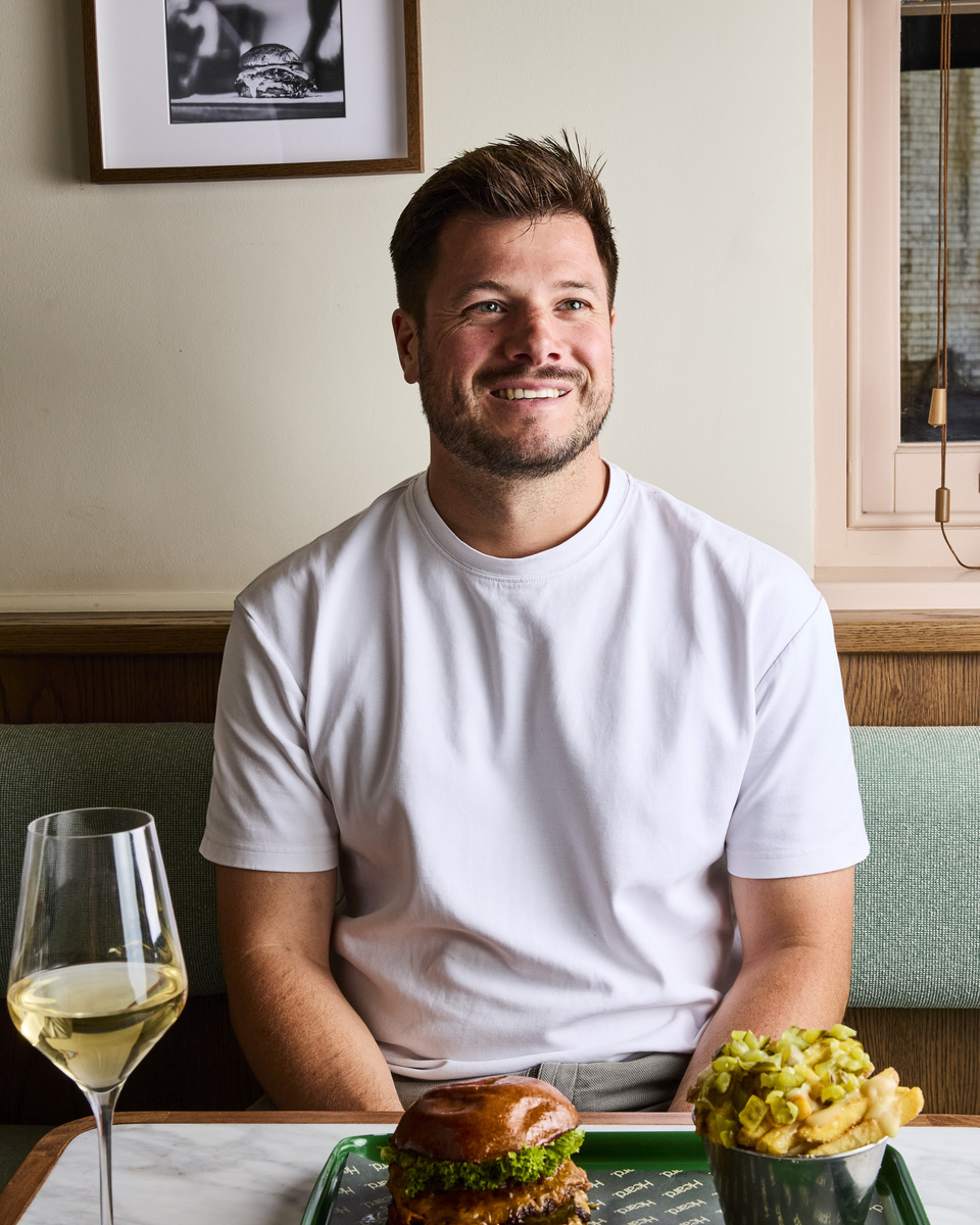 Chef Jordan Bailey sat at a table with a glass of wine, a burger, and fries