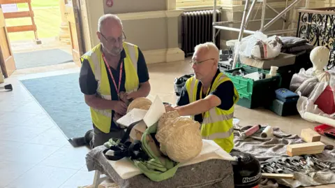 Stowe House Preservation Trust Two men in high-vis jackets working on a bust which is lying flat on a small table in a large room. They are surrounded by tools and scaffolding. 