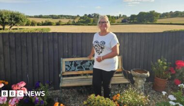 A woman with blonde hair in a ponytail and black glasses is smiling. She is standing in front of a wooden bench, which is in front of a fence - beyond which are an expanse of farmland fields and trees. In the garden in front of here are flowers and plants, as well as a bird bath