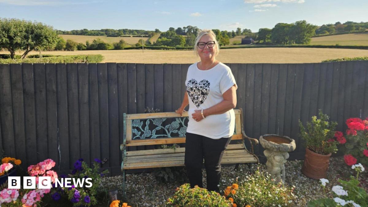 A woman with blonde hair in a ponytail and black glasses is smiling. She is standing in front of a wooden bench, which is in front of a fence - beyond which are an expanse of farmland fields and trees. In the garden in front of here are flowers and plants, as well as a bird bath