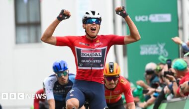 A cyclist with his arms aloft as he crosses the line first. He is wearing a red top and a cycling helmet and wrap around sunglasses. He is being followed by other cyclists and there is a crowd of people on the right hand side behind the hoardings.