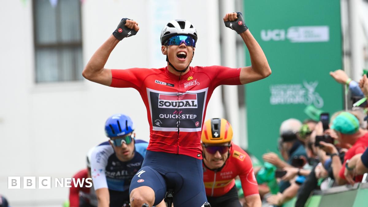 A cyclist with his arms aloft as he crosses the line first. He is wearing a red top and a cycling helmet and wrap around sunglasses. He is being followed by other cyclists and there is a crowd of people on the right hand side behind the hoardings.