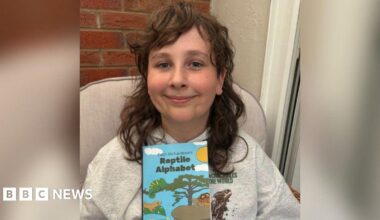 A young boy with long brown hair is smiling into the camera in a head and shoulders shot. He is wearing a grey hoodie with a brown reptile on it and holding up a blue children's book called 'Zach Richardson's Reptile Alphabet' with pictures of reptiles and a tree, sky and sun.