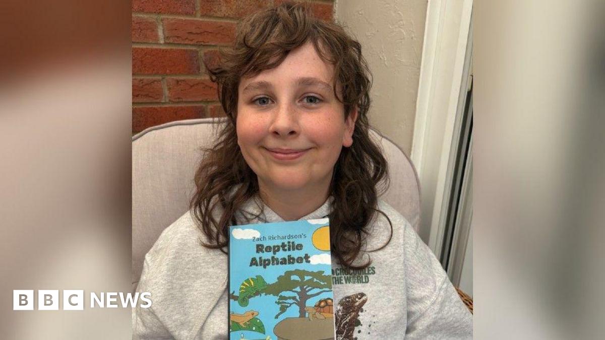 A young boy with long brown hair is smiling into the camera in a head and shoulders shot. He is wearing a grey hoodie with a brown reptile on it and holding up a blue children's book called 'Zach Richardson's Reptile Alphabet' with pictures of reptiles and a tree, sky and sun.