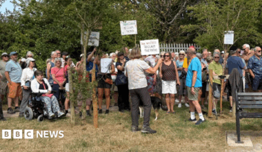 A group of protestors stand on the grass by trees. They are being spoken to by an organiser. Some protestors hold signs that say 'Dog walkers reclaim the path'.