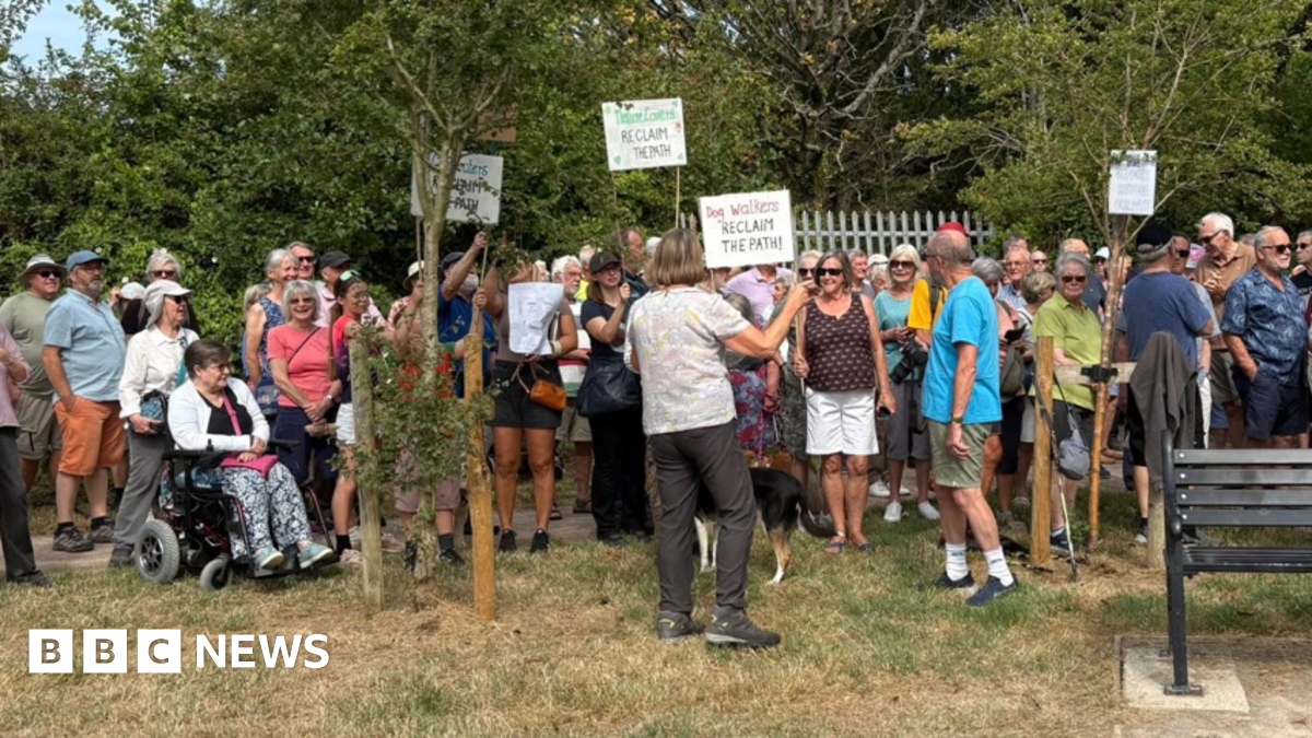 A group of protestors stand on the grass by trees. They are being spoken to by an organiser. Some protestors hold signs that say 'Dog walkers reclaim the path'.