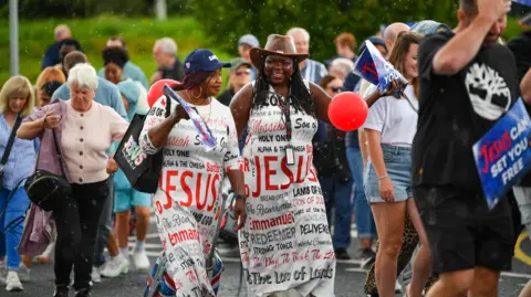Pacemaker A group of people march down a road. Two ladies are dressed in white dresses which say words like Jesus and Redeemer on it. 