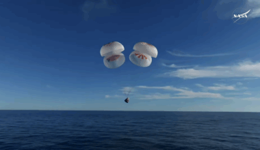 a white space capsule descends toward the ocean under four white and red parachutes