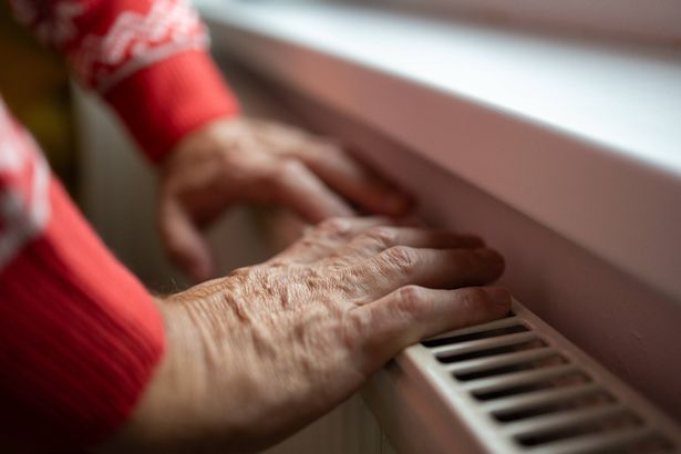 Stock image of a senior man turning the battery heating knob and warming himself from the radiator. Heating in an apartment, at home. heating season and price crises