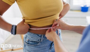 Stock image of a woman in a yellow top and jeans having her waist measured.