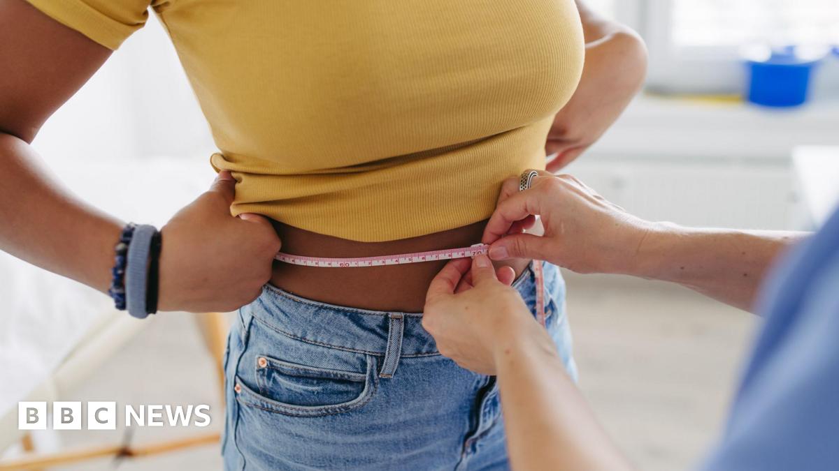 Stock image of a woman in a yellow top and jeans having her waist measured.