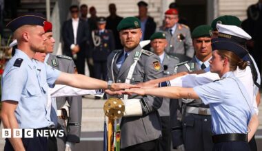 German army recruits take an oath, dressed in light blue shirts and dark blue berets as officers look on