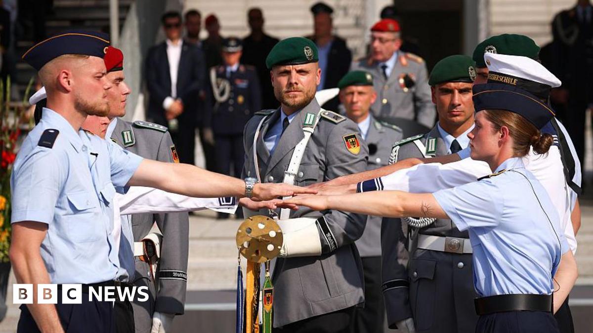 German army recruits take an oath, dressed in light blue shirts and dark blue berets as officers look on