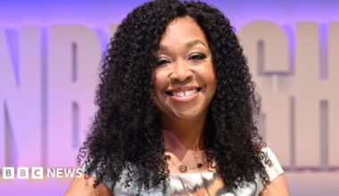 Shonda Rhimes, with a bright sile and dark curls, holds her award sitting on set at her TV festival show. The background is pale purple and she wears a patterned white, orange and black dress while sitting on a white chair.