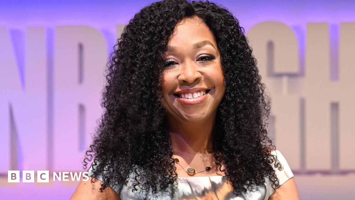 Shonda Rhimes, with a bright sile and dark curls, holds her award sitting on set at her TV festival show. The background is pale purple and she wears a patterned white, orange and black dress while sitting on a white chair.